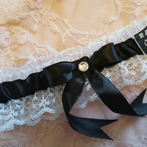 Könnte beinhalten: Ein weißes Spitzen-Strumpfband mit einem schwarzen Satinband und Schleife. Ein schwarzes und silbernes Raiders-Logo ist am Strumpfband befestigt.
