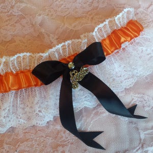 May include: A white lace garter with an orange satin ribbon and a black bow. A gold charm with a key design is attached to the bow.