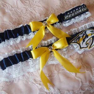 May include: Two white lace garters with navy blue satin ribbon and yellow bows. The top garter has a black patch with the text "Nashville Predators". The bottom garter has a black and yellow patch with a predator logo.