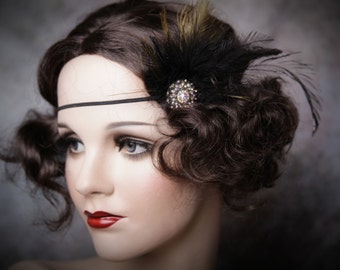 May include: A vintage-style headband featuring a black elastic band and a decorative cluster of black and gold feathers. A sparkling, round, rhinestone embellishment adds a touch of glamour. The mannequin has dark brown hair and red lipstick.