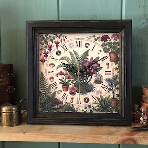 May include: A square wall clock with a dark distressed wooden frame and a cream-coloured face featuring Roman numerals. The clock face is decorated with botanical illustrations of ferns, orchids, fuchsia flowers, mushrooms, and a small hedgehog.