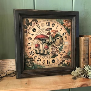 Op de afbeelding: Een vierkante wandklok met een donkerbruine houten lijst en een crèmekleurige wijzerplaat met een botanische illustratie van diverse paddenstoelen en mos. De klok heeft zwarte Romeinse cijfers en twee zwarte metalen wijzers.