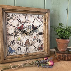 May include: A square wall clock with a weathered wooden frame and a cream-coloured face featuring Roman numerals, illustrations of various butterflies, ivy vines, and the text The Garden Hours in the centre, with black ornate clock hands.