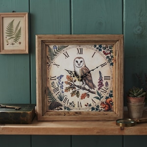 May include: A square wooden wall clock featuring a barn owl illustration at the center, surrounded by botanical and woodland animal motifs. The clock face displays black Roman numerals and ornate black hands against a cream-colored background.