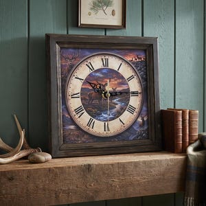 May include: A square wall clock with a dark brown wooden frame and a circular face featuring Roman numerals. The clock face displays a detailed illustration of deer in a mountainous landscape at sunset, with black metal hands indicating the time.