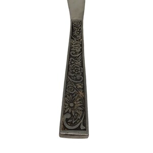 May include: A silver butter knife with a floral design etched into the handle. The handle is rectangular with a detailed pattern of flowers and leaves. The blade is smooth and rounded.