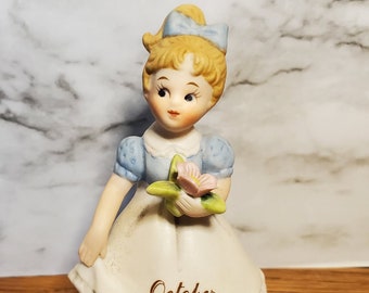 May include: A porcelain figurine of a young girl in a blue dress holding a pink flower. The figurine has blonde hair and a blue bow. The word "October" is printed on the apron. The figurine is approximately 5 inches tall.
