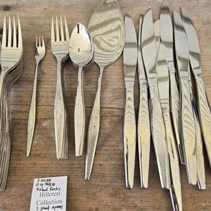 Könnte beinhalten: Eine Sammlung von Vintage-Besteck, darunter Gabeln, Messer, Löffel und Servierutensilien. Das Set ist auf einer Holzoberfläche angeordnet. Der handgeschriebene Zettel weist auf die Hillcrest-Kollektion hin.