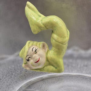 Pode incluir: Uma pequena estatueta de cerâmica verde-clara de um elfo numa pose arqueada e contorcida. O elfo tem um rosto pintado com olhos pretos, sobrancelhas arqueadas e boca vermelha, usando um gorro pontiagudo e um fato verde com acabamento salpicado.