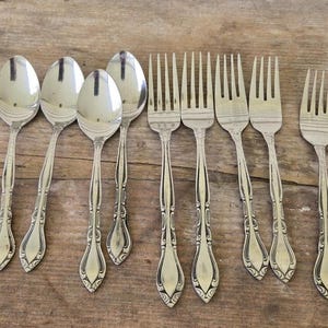 Könnte beinhalten: Eine Sammlung von Vintage-Besteck, darunter Löffel und Gabeln, angeordnet auf einer rustikalen Holzoberfläche. Das Besteck hat verzierte Griffdetails und eine polierte Oberfläche, was auf eine klassische oder antike Ästhetik hindeutet.