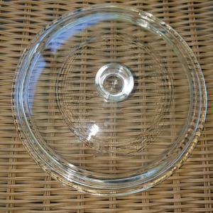 May include: A clear glass lid with a circular design and a small, round handle in the center. The lid has a slightly raised edge and is designed to fit on a pot or pan. The glass is transparent, allowing visibility of the contents.