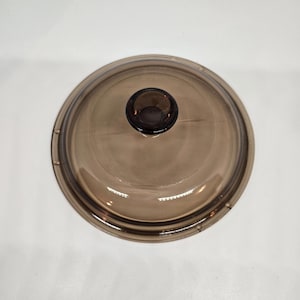 Puede incluir: Una tapa de vidrio transparente y redonda con un pomo marrón oscuro. La tapa está ligeramente teñida y parece ser para una olla o sartén. El pomo también es marrón oscuro y redondo. La tapa está sobre una superficie blanca.