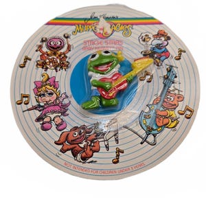 May include: A Jim Henson's Muppet Babies Stage Stars Baby Kermit pin on a circular cardboard backing. The pin features a green frog playing a red guitar. The backing displays illustrations of Muppet characters, musical notes, and the text NOT INTENDED FOR CHILDREN UNDER 3 YEARS.
