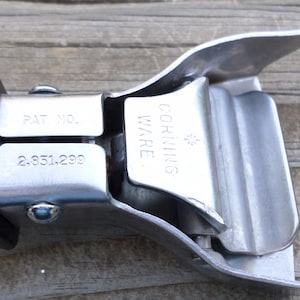 Può includere: Primo piano di una clip per manico di pentola Corning Ware. La clip è realizzata in metallo argentato e plastica nera. La clip riporta la scritta "CORNING WARE" e "PAT NO. 2.851.299".