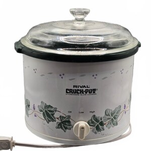 May include: A white Rival Crock-Pot stoneware slow cooker with a clear glass lid. The exterior features a green ivy leaf and purple berry vine pattern. It has a front-facing control knob with Off, Low, and High settings and a white power cord.
