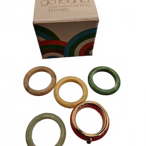 May include: A set of five rings and a cardboard box labeled Avon color go round convertible ring (small). The rings include a marbled tan band, a solid yellow band, a green band, a pale green band, and a red band with a gold-tone metal rim.