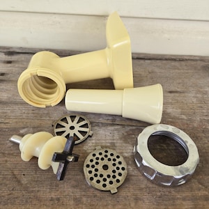 Op de afbeelding: Een assortiment vintage roomkleurige vleesmolenonderdelen, waaronder de hoofdbehuizing, een toevoerbuis, een wormschroef en diverse maalplaten. De onderdelen zijn gerangschikt op een verweerd houten oppervlak.