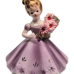 May include: A vintage porcelain figurine of a young woman in a lavender dress, holding a basket of pink flowers. The figurine has a detailed face, with blonde hair and gold accents on the dress. The dress is decorated with floral and leaf designs.