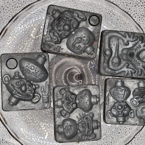 May include: Five rectangular gray metal molds featuring embossed cartoon character designs, including a figure with the text 'JOY' and another with 'ELVIS'. A wooden ruler is positioned on the left, showing the molds are approximately 3 inches wide.