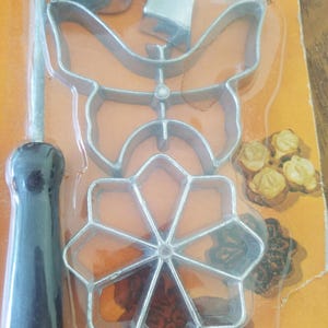 May include: A vintage set of metal cookie cutters in various shapes, including a butterfly and a flower, packaged in clear plastic. The set includes a black-handled tool. The packaging is orange and yellow.