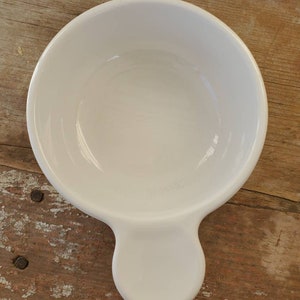May include: A white ceramic bowl with a handle. The bowl is round with a smooth, glossy finish. The handle is a rounded shape, extending from the side of the bowl. The bowl is sitting on a weathered wooden surface.