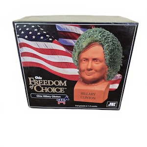 May include: A Chia Hillary Clinton planter with a terracotta bust and green chia hair, packaged in a box with an American flag background. The box reads "Freedom of Choice" and "Chia Hillary Clinton."
