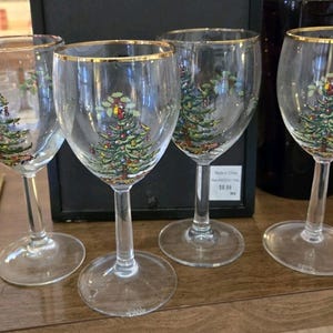 May include: Set of four clear wine glasses with gold rims. Each glass features a painted Christmas tree design with colorful ornaments. The glasses are displayed on a wooden surface, suggesting a festive holiday theme.