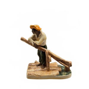 Può includere: Una statuetta in ceramica raffigurante un personaggio con cappello giallo, camicia chiara e pantaloni scuri, appoggiato a un lungo tronco di legno. Il personaggio poggia su una base composta da diversi tronchi con una piccola ascia a lato.