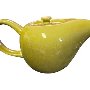 May include: A yellow-green ceramic gravy boat with a curved handle and a spout. The gravy boat has a lid with a small knob and a crackle glaze finish. The design is smooth and rounded, suitable for serving sauces.