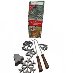 May include: A 7-piece holiday rosette iron set including a cardboard box labeled SKU 1017577 and 7 Piece Holiday Rosette Iron Set. The set features metal molds in shapes of an angel, star, bell, snowflake, and tree, along with two metal handles with wooden grips.