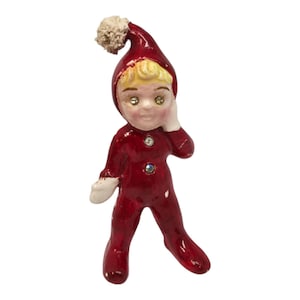 May include: A vintage ceramic figurine of a child dressed in a red elf suit with a matching hat and a white pom-pom. The figurine has blonde hair, a fair complexion, and rhinestone eyes and buttons. The figure is posed with one hand near its face.