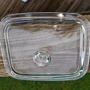 May include: A clear, rectangular glass lid with rounded corners and a circular handle. The lid is designed to fit a rectangular dish. The glass is transparent, allowing a view of the contents below.