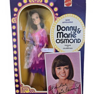 May include: A vintage Mattel doll of Marie Osmond in a box. The doll is wearing a pink and purple dress and holding a microphone. The box features the text "Donny & Marie Osmond" and a photo of Marie Osmond.