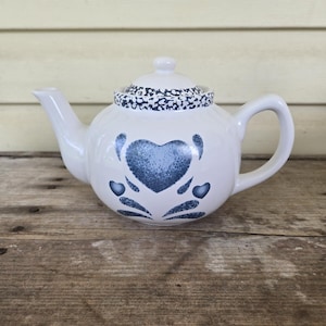 Op de afbeelding: Witte keramische theepot met een blauw hartontwerp en bijpassend deksel. De theepot heeft een afgeronde body, een gebogen handvat en een tuit. Het deksel is versierd met een blauw-wit gespikkeld patroon. De theepot staat op een houten oppervlak.