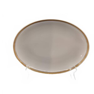 May include: Oval-shaped, off-white ceramic plate with a gold-colored rim and a thin black accent line. The plate is displayed on a clear stand against a white background. A simple, elegant design for serving food.