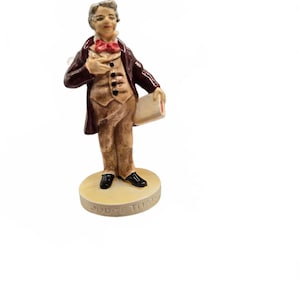 Può includere: Una statuetta in ceramica dipinta a mano raffigurante una persona con cappotto marrone, gilet color cuoio e papillon rosso, che tiene in mano un libro bianco. La figura poggia su una base circolare con il testo in rilievo JUDGE THAT.