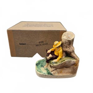 Può includere: Una statuetta in ceramica di un personaggio con cappello e camicia gialli e pantaloni scuri, seduto su una roccia accanto a un tronco d'albero. La base reca l'iscrizione HUCKLEBERRY FINN. Dietro c'è una scatola di cartone etichettata 6133 HUCK FINN.