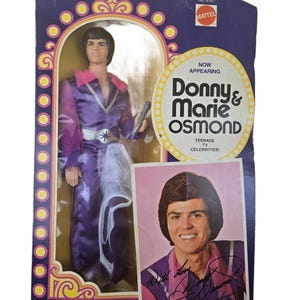 May include: A vintage Donny & Marie Osmond doll in its original packaging. The doll is dressed in a purple jumpsuit with a silver belt and holds a microphone. The box features the text "Donny & Marie Osmond" and a photo of Donny Osmond.