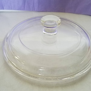May include: A clear glass lid with a small, rounded knob. The lid is circular and appears to be for a pot or pan. The glass is transparent, allowing a view of the contents below. The knob is also clear.