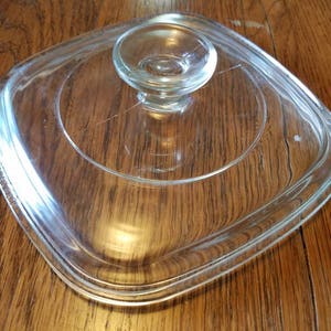 May include: A clear, square glass lid with a rounded handle. The lid has a slightly raised rim and is designed to fit a square pot or pan. The handle is also clear and rounded, providing a secure grip.