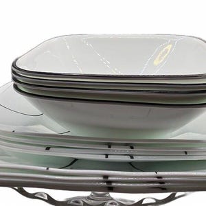 May include: A stack of white square dinnerware featuring a thin black border and subtle black line accents. The set includes multiple square plates and deep square bowls, all arranged in a neat vertical pile against a plain white background.