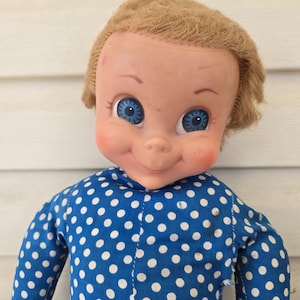 May include: A doll with a molded plastic head, light brown hair, and bright blue eyes. The doll has a smiling expression with rosy cheeks and is wearing a long-sleeved blue shirt featuring a repeating pattern of small white polka dots.