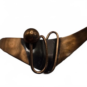 Op de afbeelding: Een broche met een donkere, gebogen, driehoekige metalen basis met een gepolijste bronzen afwerking. Het item is voorzien van een kleine, bolvormige bronzen kraal en twee langwerpige, ovaalvormige draadlussen die over het donkere middendeel vallen.