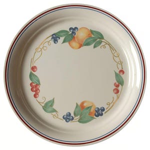 May include: A round cream-colored ceramic plate featuring a central decorative wreath of painted fruit including oranges, blueberries, and red berries with green leaves and gold vines. The rim is accented with thin concentric red and blue circular lines.