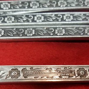 Puede incluir: Primer plano de cubiertos de acero inoxidable con grabados florales y de hojas. Los mangos están decorados con un patrón repetido de flores y hojas. El texto "Gold Standard Stainless Japan A" es visible.