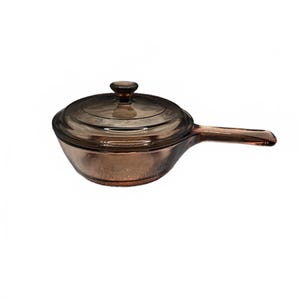 May include: A translucent brown glass saucepan with a matching lid. The round pan features a single extended handle on the side and a circular knob on the centre of the lid. The glass has a smooth, reflective surface and a deep amber tint throughout.