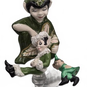 May include: A ceramic figurine featuring a large elf in a dark green outfit with a pointed hat, holding a smaller elf dressed in green with white fuzzy trim. A third, smaller elf figure in a green outfit and black boots is positioned to the side.