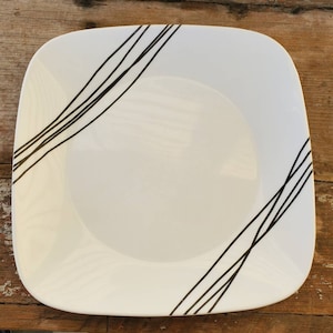 Puede incluir: Plato blanco cuadrado con esquinas redondeadas, decorado con líneas diagonales negras. Las líneas son finas y espaciadas, creando un diseño moderno. El plato es probablemente de cerámica o porcelana, ideal para servir alimentos.
