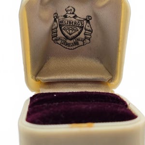Pode incluir: Uma caixa de joias aberta na cor creme com um encaixe para anel em veludo roxo escuro. O interior da tampa possui um forro de cetim creme estampado com um logotipo preto da Helzberg's Quality Value Jewelers com um diamante.