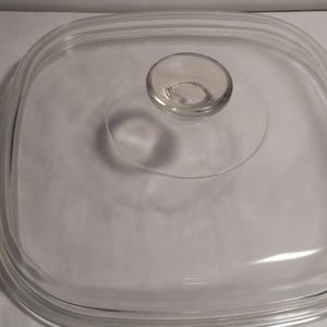 May include: A clear, square glass lid with rounded corners and a small, round handle. The lid has a slightly raised rim and is designed to fit a square cooking dish. The glass is transparent.